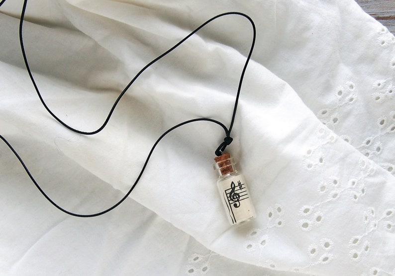 Puede incluir: Un collar con un cord&oacute;n negro y un peque&ntilde;o colgante de botella de vidrio. La botella contiene un trozo de papel con una clave de sol y notas musicales impresas en &eacute;l.