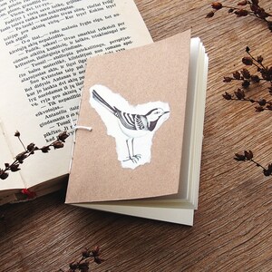 May include: A small brown notebook with a white bird illustration on the front cover. The notebook is closed and has a white string tied around it.