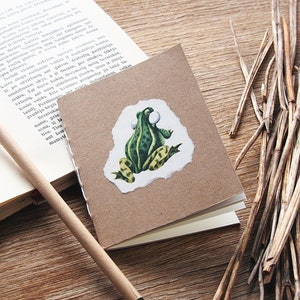 Könnte beinhalten: Ein kleines braunes Notizbuch mit einer grünen Frosch-Illustration auf dem vorderen Umschlag. Der Frosch sitzt auf einem Stück Papier mit einem zerrissenen Rand. Das Notizbuch liegt auf einer Holzoberfläche.