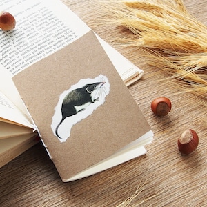 May include: A small brown notebook with a white page turned open. A brown and white illustration of a shrew is glued to the front cover. The notebook is sitting on a wooden surface with wheat stalks and hazelnuts.