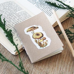 May include: A small brown notebook with a mushroom illustration on the front cover. The notebook is lying on a wooden surface with a pencil and green sprigs.
