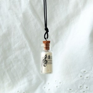Puede incluir: Un collar con una botella de vidrio y un tap&oacute;n de corcho. La botella contiene un trozo de papel con una clave de sol y notas musicales impresas. El collar est&aacute; en un cord&oacute;n negro.