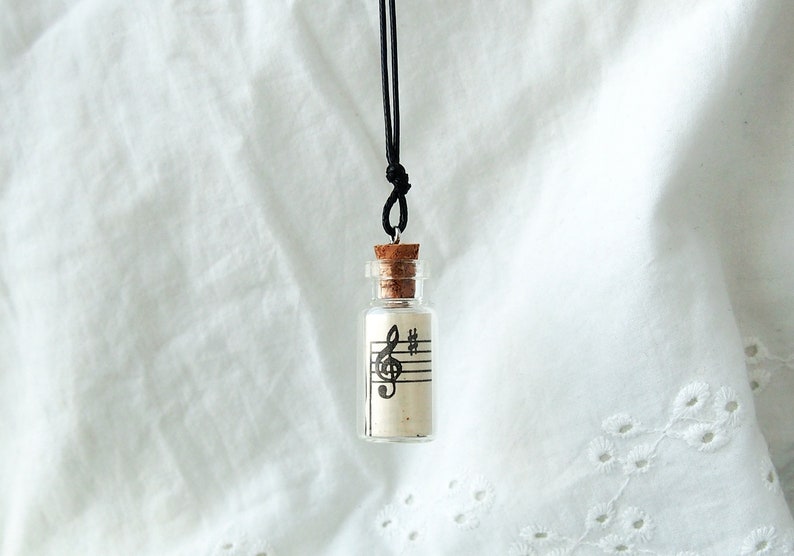 Puede incluir: Un collar con una botella de vidrio y un tap&oacute;n de corcho. La botella contiene un trozo de papel con una clave de sol y notas musicales impresas. El collar est&aacute; en un cord&oacute;n negro.