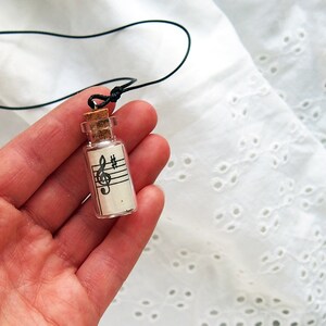 Puede incluir: Una peque&ntilde;a botella de vidrio con un tap&oacute;n de corcho, que contiene un trozo de papel con una clave de sol y notas musicales impresas en &eacute;l. La botella est&aacute; unida a un cord&oacute;n negro, lo que la convierte en un collar.