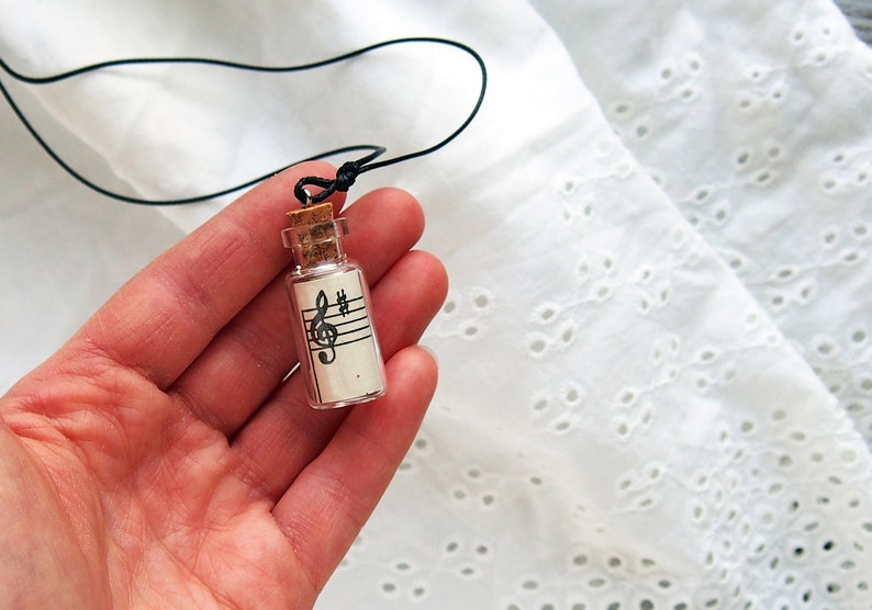 Puede incluir: Una peque&ntilde;a botella de vidrio con un tap&oacute;n de corcho, que contiene un trozo de papel con una clave de sol y notas musicales impresas en &eacute;l. La botella est&aacute; unida a un cord&oacute;n negro, lo que la convierte en un collar.
