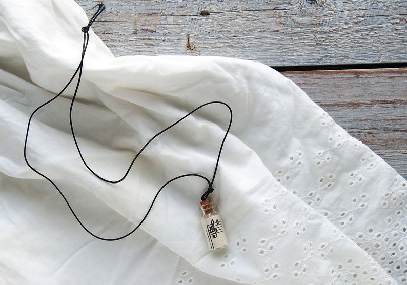 Puede incluir: Un collar de cord&oacute;n negro con un peque&ntilde;o colgante de botella de vidrio. La botella contiene un trozo de papel con una nota musical impresa.