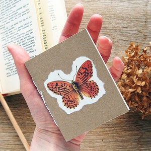 May include: A small brown notebook with a torn paper edge, featuring a vintage illustration of a red and orange butterfly.