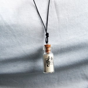 Puede incluir: Un collar con un cord&oacute;n negro y un peque&ntilde;o colgante de botella de vidrio. La botella contiene un trozo de papel con una clave de sol impresa.