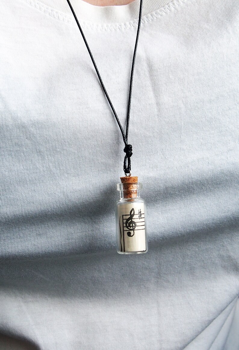 Puede incluir: Un collar con un cord&oacute;n negro y un peque&ntilde;o colgante de botella de vidrio. La botella contiene un trozo de papel con una clave de sol impresa.