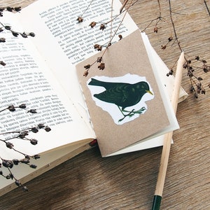 May include: A brown paper card with a black bird illustration on it. The card is sitting on top of an open book with a pencil next to it. The book is on a wooden surface with dried branches.
