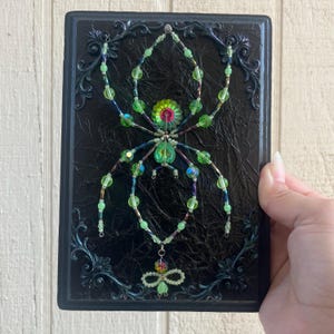 May include: A rectangular black plaque with an ornate border featuring a central spider figure crafted from light green and iridescent beads. The spider has a flower-shaped center and long, multi-beaded legs, with a small beaded charm hanging below.