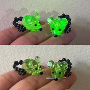 Puede incluir: Dos pares de pendientes de ratón de cristal verde. Cada ratón tiene una cola de cuentas negras, ojos de cuentas negras y bigotes plateados. Los pendientes se sostienen en una mano.