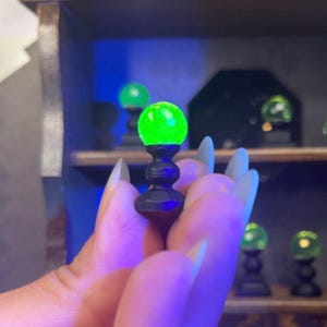 Puede incluir: Una pequeña esfera verde brillante se asienta sobre un soporte negro y ornamentado. La esfera se sostiene en una mano, con otras esferas similares visibles en el fondo. Las esferas parecen ser parte de una colección o exhibición, posiblemente para una casa de muñecas o un entorno de fantasía.