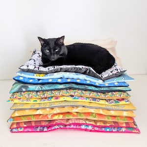 May include: A black cat resting on a stack of rectangular fabric pet mats. The mats feature various colourful patterns including floral designs, citrus fruit prints, and sugar skull motifs in shades of pink, yellow, orange, blue, and green.