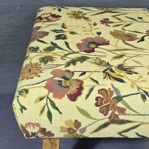 May include: A rectangular ottoman featuring a cream-coloured fabric upholstery with a woven floral pattern in shades of red, pink, yellow, green, and dark blue. The piece is supported by light-toned wooden legs visible at the bottom corners.