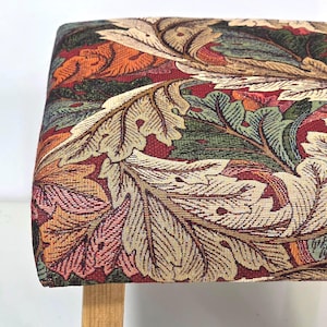 May include: A rectangular stool featuring a woven tapestry fabric seat with a dense pattern of large, stylised leaves in shades of deep red, forest green, golden yellow, and tan. The stool is supported by light-coloured wooden legs.