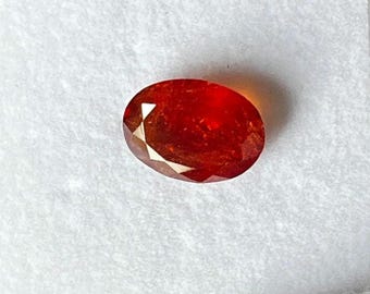 May include: Oval-shaped, faceted gemstone with a vibrant red-orange hue. The gem displays a polished surface with multiple facets, reflecting light. The gemstone is set against a white background, highlighting its color and clarity. A close-up view showcases the gem's intricate cut.