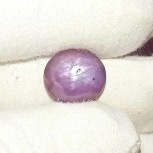 Può includere: Una gemma sferica, lucidata, rosa-violacea, possibilmente un rubino stellato, è appoggiata su un tessuto bianco. La gemma ha una superficie liscia con sottili riflessi di luce. Il colore della pietra è un rosa tenue e smorzato con un pizzico di lavanda.