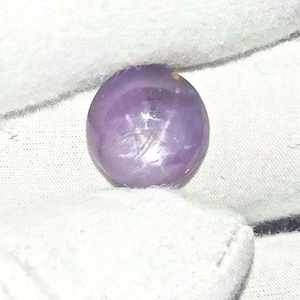 May include: A round, polished gemstone with a soft, lavender hue. The gem has a smooth surface with subtle internal patterns and is set against a white background.
