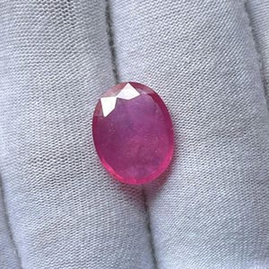 May include: Oval-cut ruby gemstone with a deep pink hue. The gemstone has a faceted top, reflecting light. The ruby is set against a white fabric background, highlighting its color and clarity. A precious stone for jewelry.