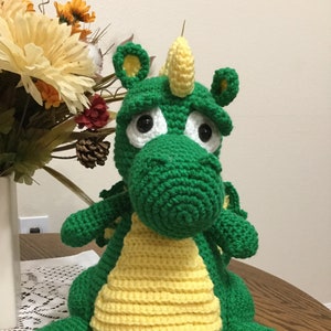 May include: A green crocheted dragon with yellow accents and large black eyes. The dragon is sitting on a white crocheted doily.