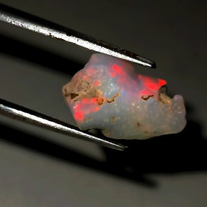 Puede incluir: Un ópalo en bruto translúcido con una base de color blanco lechoso y destellos de color rojo intenso en toda su superficie irregular y texturizada, sostenido firmemente por unas pinzas de metal plateado sobre un fondo oscuro.