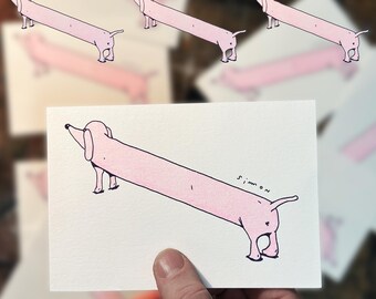 May include: A pink dachshund with a long body and short legs is drawn on a white card. The card is held in a person's hand. The text "simmon" is written in black ink on the card.