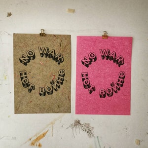 Könnte beinhalten: Zwei Poster mit dem Text "No War, Not Here, Bow Wow He" in Schwarz auf braunem und rosa Hintergrund.