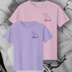 Könnte beinhalten: Zwei kurzärmelige T-Shirts, eines in Helllila und eines in Rosa, werden gezeigt. Jedes Shirt hat eine kleine, stilisierte Hunde-Grafik auf der Brust. Der Hintergrund ist ein hellgrauer Stoff mit einer sich wiederholenden Hunde-Illustration.