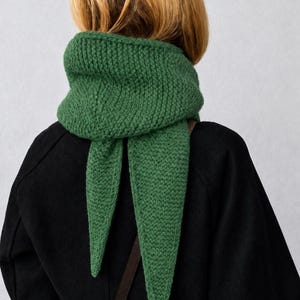May include: A forest green knitted scarf with a triangular shape and a textured garter stitch pattern. The scarf is wrapped around the neck, featuring two pointed ends that hang down the back over a solid black coat with a brown strap.