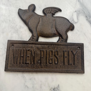 Op de afbeelding: Een gietijzeren bord met een varken met vleugels, boven een rechthoekige plaat. De plaat leest "WHEN PIGS FLY" in verhoogde letters. Het bord heeft een rustieke, bruine afwerking.