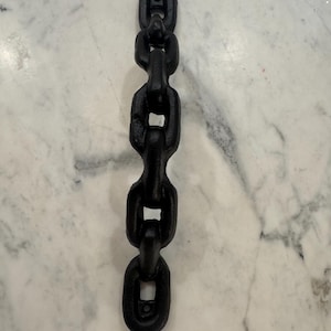 May include: A vertical, matte black chain with five interlocking links. The chain is made of a solid material and has a slightly textured surface. The links are oval-shaped and connected in a repeating pattern. The chain is displayed against a light-colored background.