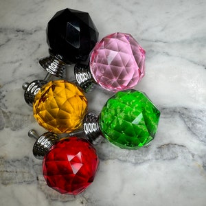 May include: Five crystal cabinet knobs in various colors, including black, pink, yellow, green, and red. Each knob has a faceted design and a silver metal base.