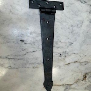 May include: Black metal T-hinge with a pointed tip. The hinge has a rectangular top plate and a long, narrow arm.