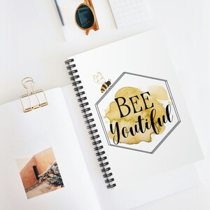 Puede incluir: Una libreta de espiral blanca con un diseño de abeja acuarela amarilla. El diseño presenta una abeja y el texto "BEE youtiful".