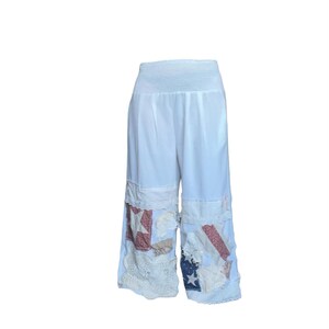 May include: White wide-leg pants with a smocked waistband. The lower legs feature patchwork designs with red, white, and blue fabric, including star and stripe patterns. The edges are trimmed with white lace.