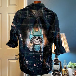 May include: A dark green and black plaid flannel shirt with a bleached effect. The shirt has a graphic of a skull wearing a cowboy hat and bandana, with the skull giving a peace sign with both hands.