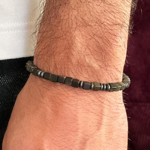 Op de afbeelding: Een donkergrijze kralenarmband met vierkante kralen en kleine zilverkleurige afstandhouders. De armband wordt om een pols gedragen, met een wit shirt en een donkere jeans op de achtergrond. Het is een sieraad.