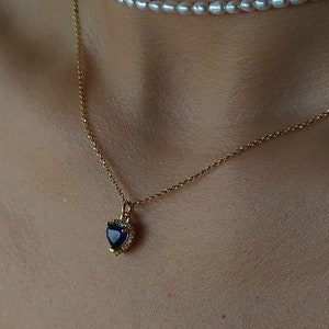 Può includere: Una collana a catena d'oro con un ciondolo a forma di cuore blu e una collana choker di perle. Le collane sono sovrapposte sul collo di una persona.