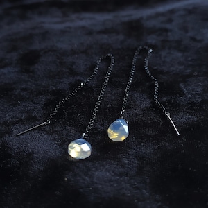 Puede incluir: Un par de pendientes colgantes con una piedra de ópalo en forma de lágrima en cada pendiente. Los pendientes tienen una cadena negra y un poste plateado.
