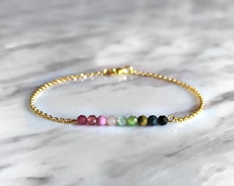 May include: A gold chain bracelet with a row of colorful gemstone beads. The beads are pink, light pink, green, gold, dark blue, and black.