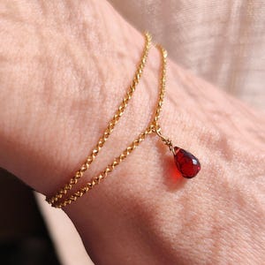 May include: A delicate gold bracelet with a double chain design. A teardrop-shaped, deep red gemstone hangs from the bracelet. The bracelet is worn on a wrist, with the skin tone visible in the background.