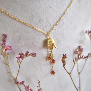 May include: A gold-colored necklace featuring a tulip-shaped pendant. The pendant has a textured surface and is adorned with dangling orange beads. The necklace is displayed against a soft, neutral background with dried flowers.