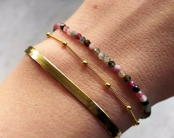 May include: A gold chain bracelet with small round beads, layered with a gold bangle bracelet and a multi-colored beaded bracelet with pink, green, and black beads.