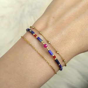 Puede incluir: Tres pulseras de oro en una muñeca. La pulsera superior es una cadena delicada con pequeñas cuentas doradas. La pulsera del medio es una pulsera de cuentas con cuentas azules, rojas y rosadas. La pulsera inferior es una cadena de oro más gruesa.