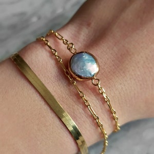 May include: A gold chain bracelet with a large, round, iridescent pearl charm. The bracelet is layered with a thin, gold, snake chain bracelet.
