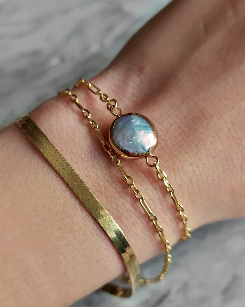 May include: A gold chain bracelet with a large, round, iridescent pearl charm. The bracelet is layered with a thin, gold, snake chain bracelet.