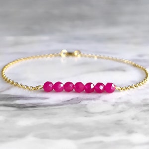May include: A delicate gold bracelet featuring a strand of small, faceted, hot pink beads. The bracelet has a delicate chain with a small clasp. The background is a light gray and white marble.