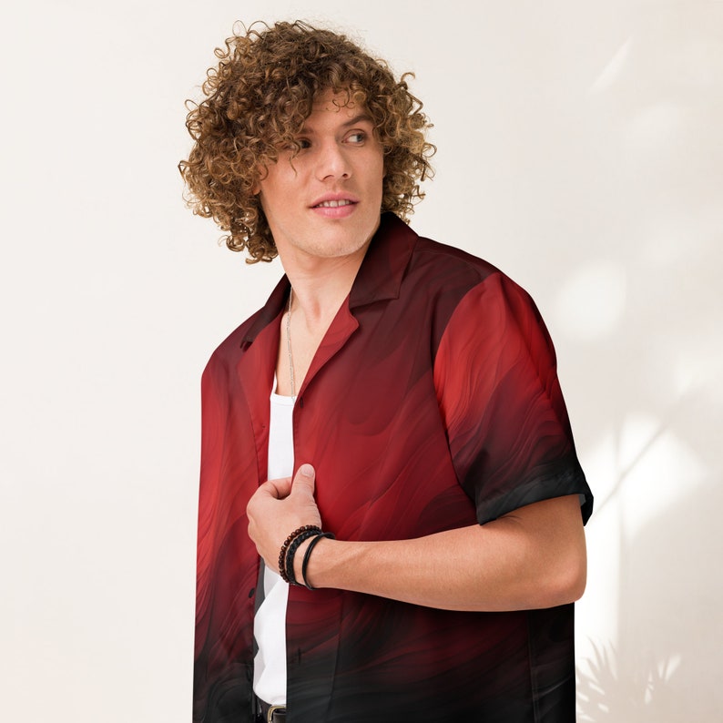 Op de afbeelding: Een man met krullend bruin haar draagt een rood en zwart, kortgeknipt overhemd met knopen en een golvend patroon. Hij draagt een zilveren kettingketting en een zwarte armband.
