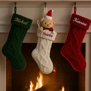 May include: Three cable-knit Christmas stockings in green, white, and red hang above a fireplace. The green stocking is embroidered with "Michael", the white with "Timothy", and the red with "Katerina". A teddy bear wearing a Santa hat peeks out of the white stocking.